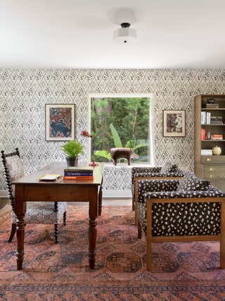 patterned wallpaper with leafy motif patterned pink rug window on far wall showing leafy plants outside wood desk...