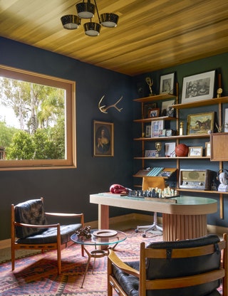 Office with dark walls window to left shelving behind desk two chairs flank a low glass table in front of desk